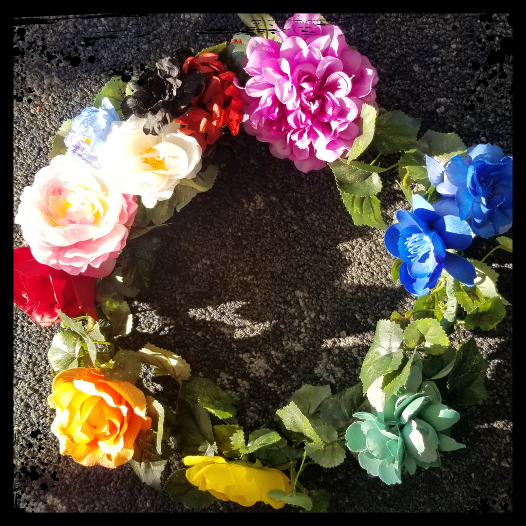 A handmade flower crown sits finished on the floor of an airport. The rug is a tough brownish color. Each flower in the crown is a color of the rainbow. At the top left of the crown, flowers in the colors brown, black, pink and blue have been added in the style of the Progress Pride Flag. The leaves are green.